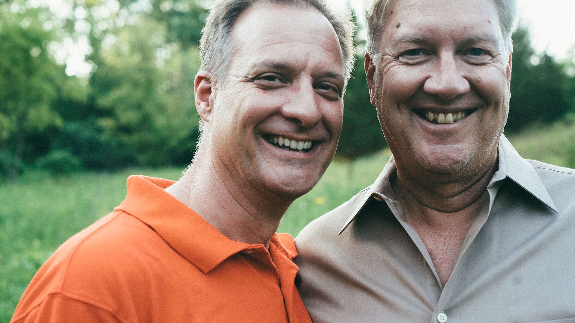 Two late-middle-aged men, standing close by and smiling cheekily to camera.